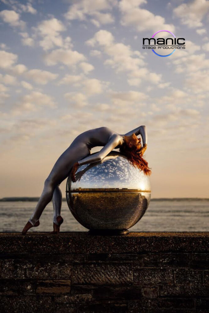 Globe contortionist dressed in silver catsuit laid over a large mirrored globe by the sea