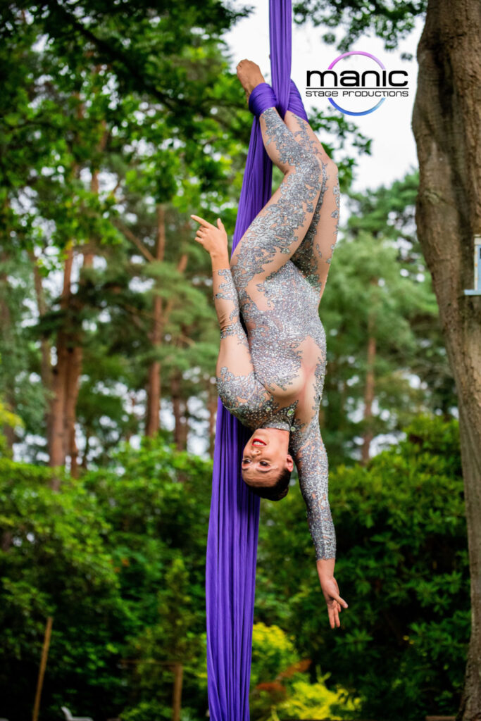 Silks aerialist hanging upside down from a set of purple silks amongst the tree tops