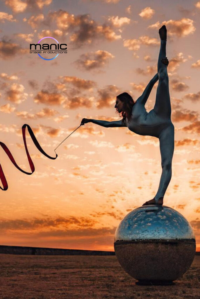 Talented gymnast and acrobat stood on a mirrored globe in the splits twirling a ribbon against the sunsetting sky