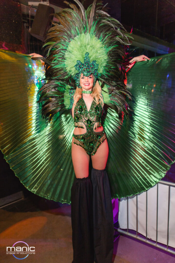 Green Vegas stilt walking showgirl holding her large green wings high above her head smiling at the camera and passing party goers